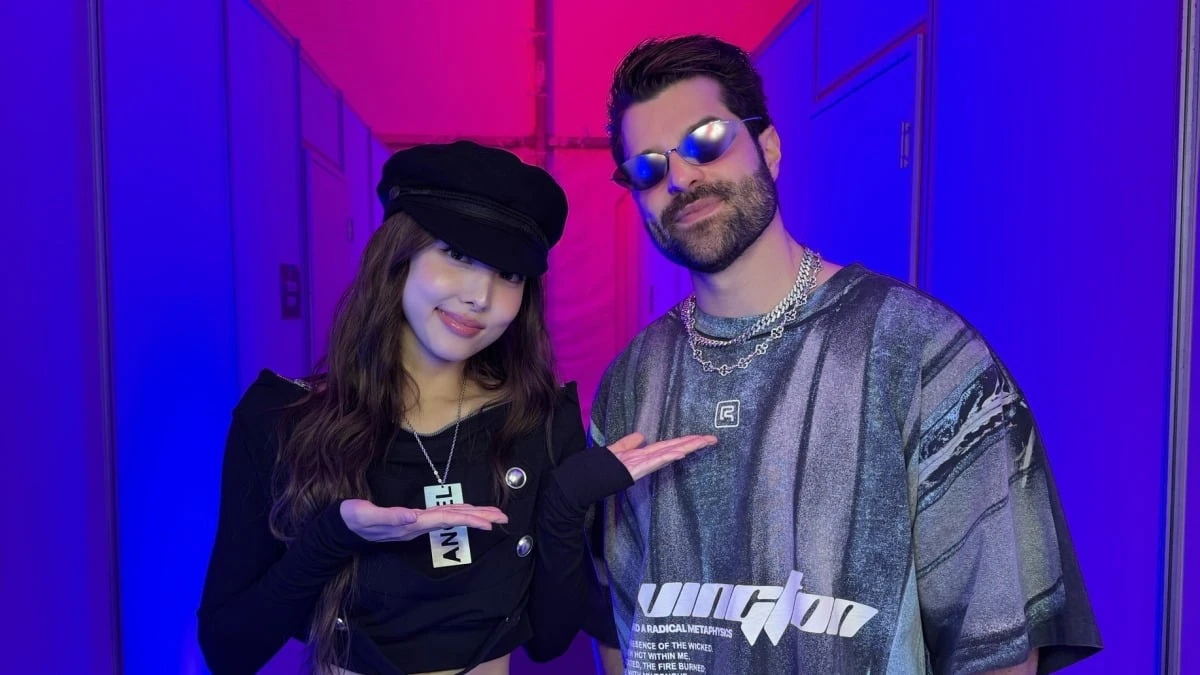 Nayeon, do TWICE, de boina preta, e Alok, de óculos espelhados e camiseta estampada, posando juntos num corredor de bastidor iluminado de azul e rosa