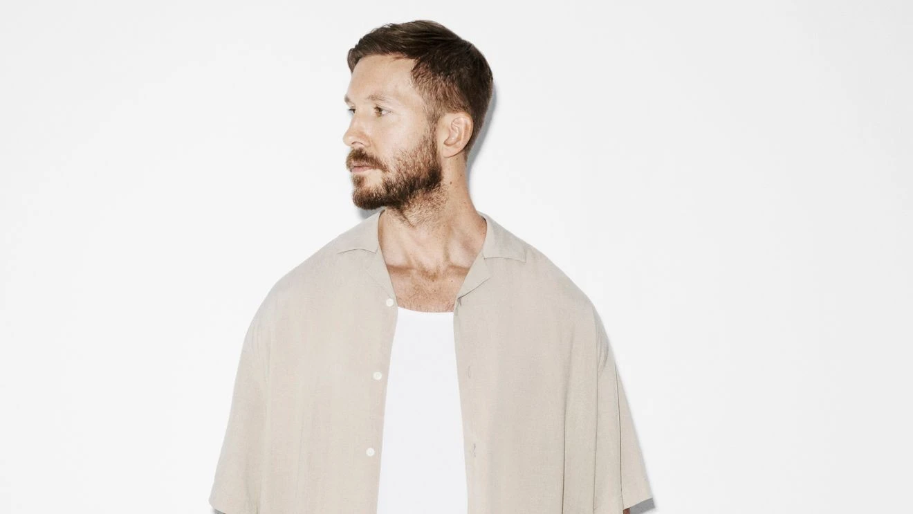 Foto de divulgação de Calvin Harris, de barba, camisa bege aberta sobre camiseta branca, olhando para o lado, contra fundo branco