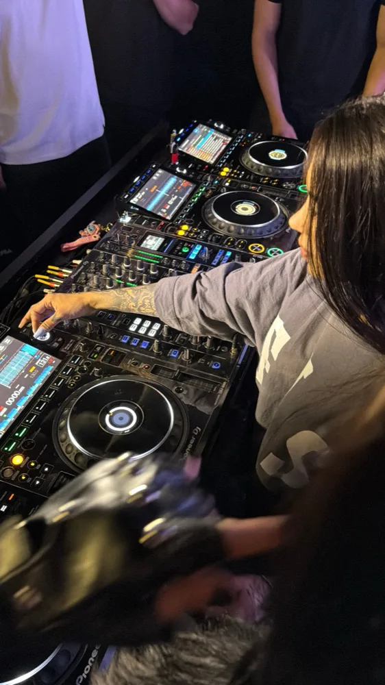 DJ mulher mixando cercada de público próximo, telas coloridas do equipamento acesas, no estúdio 7 da DJ Ban EMC