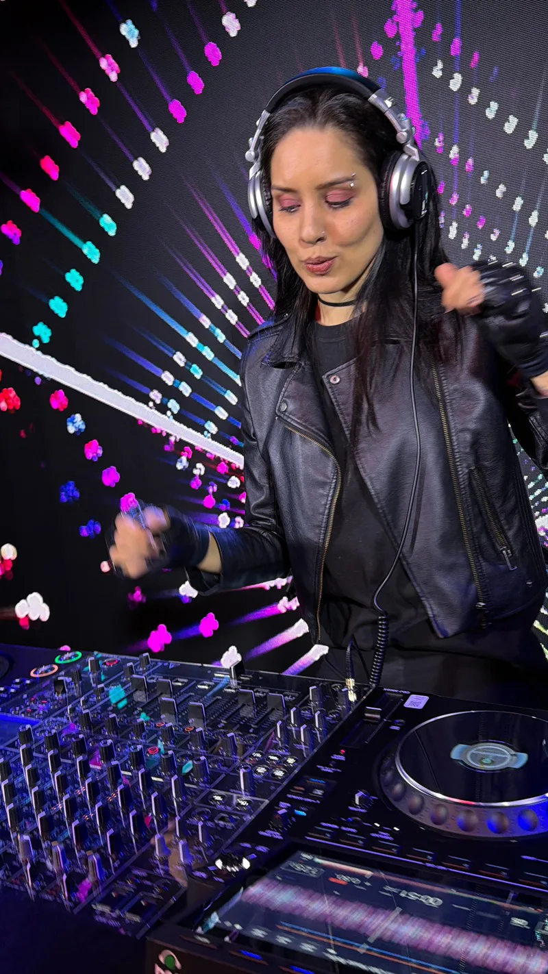 DJ mulher de jaqueta de couro tocando nos controles, com tela de LED colorida ao fundo, no estúdio da DJ Ban EMC