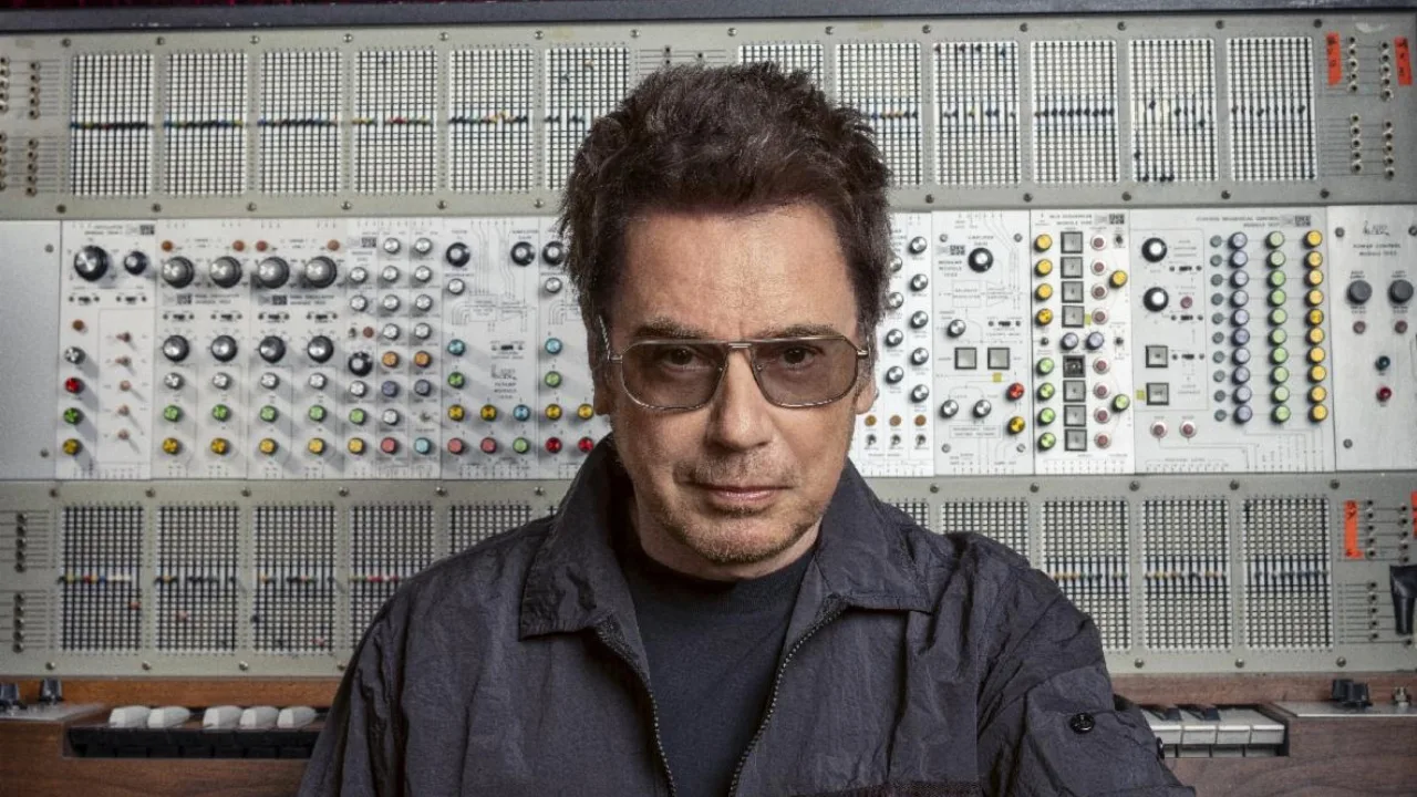 Jean-Michel Jarre de óculos de lente clara e jaqueta escura, diante de um grande sintetizador modular com dezenas de botões coloridos