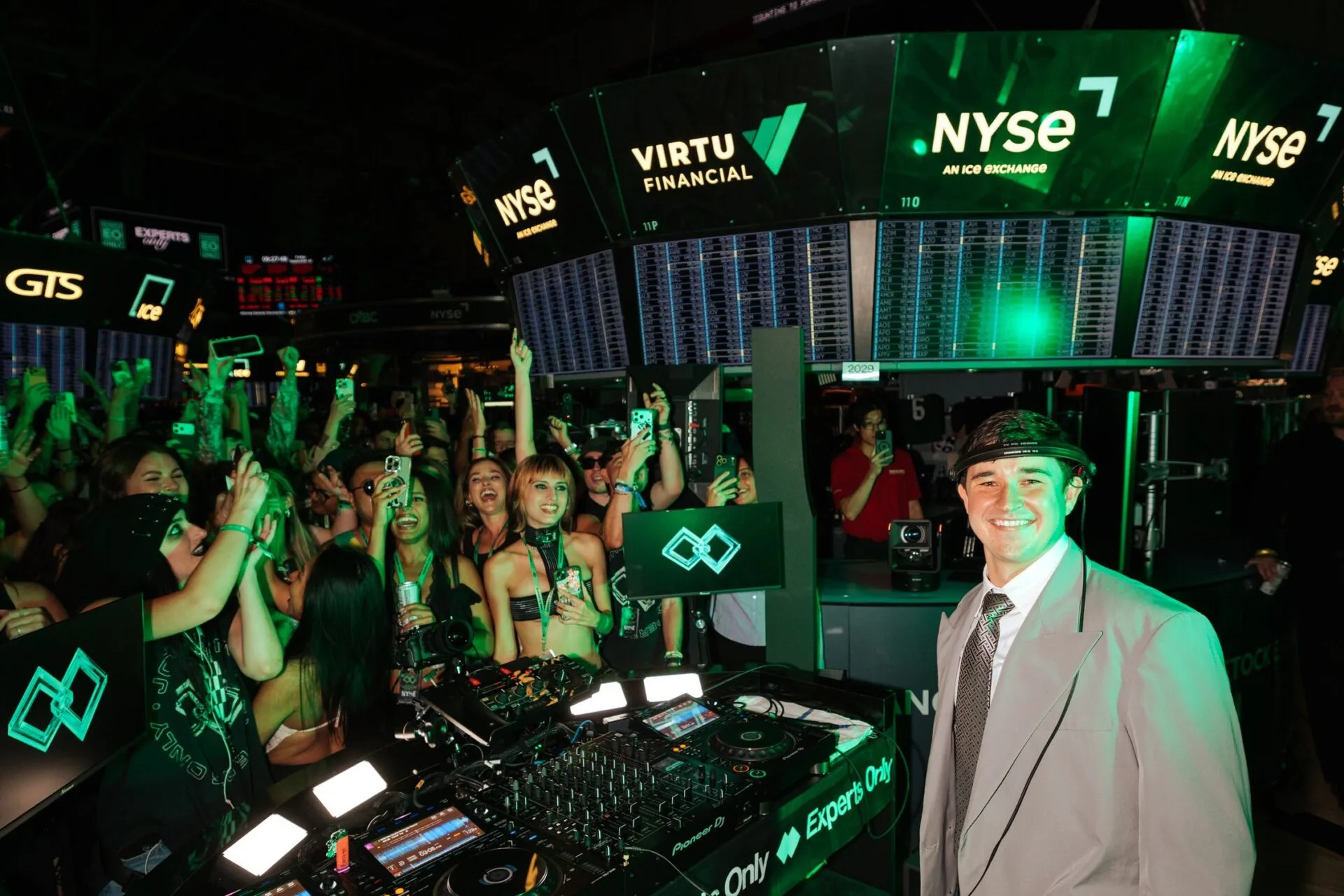 John Summit de terno cinza ao lado dos CDJs no pregão da Bolsa de Nova York, com o público e os painéis da NYSE ao fundo