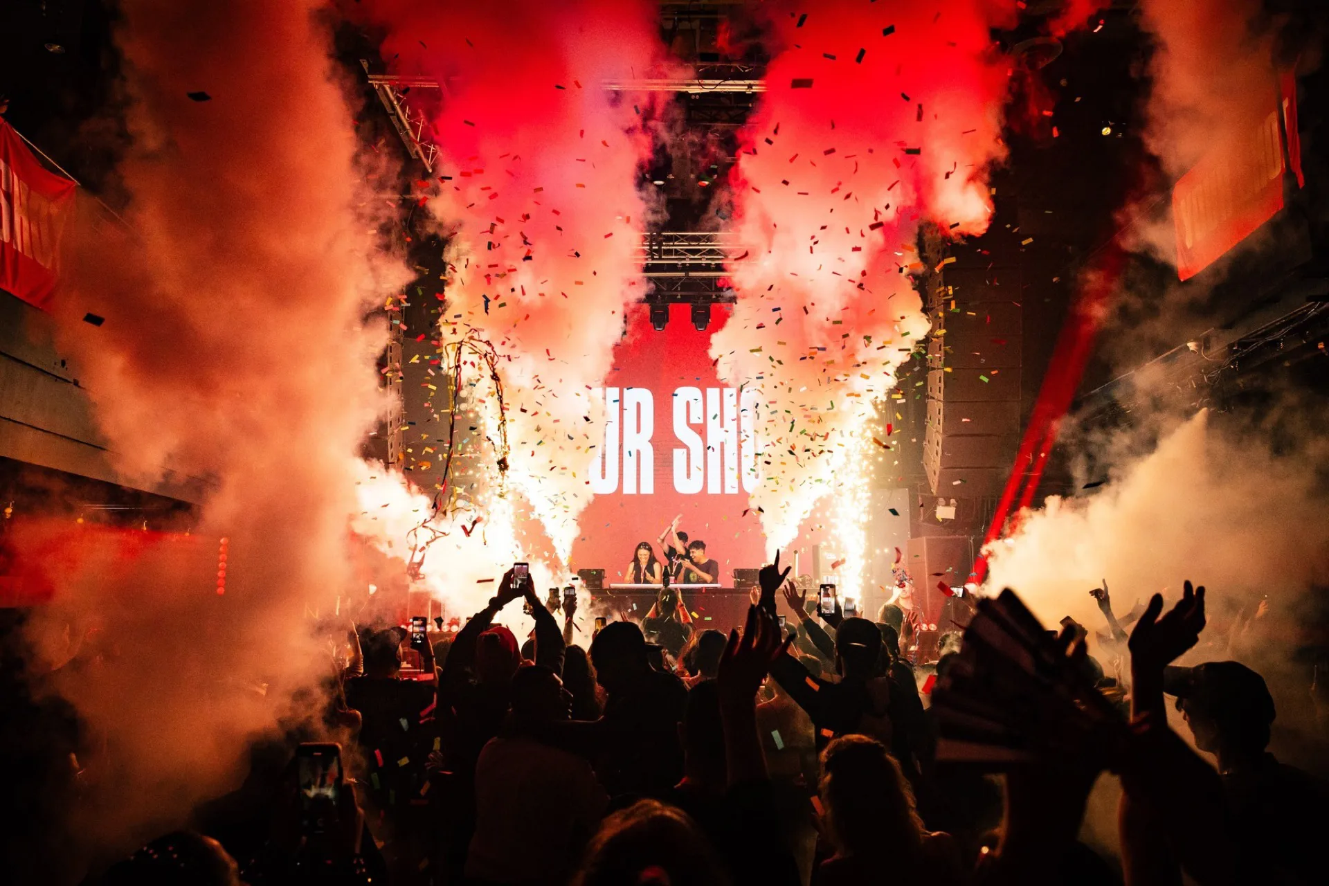Dois DJs no palco da competição Your Shot, com fumaça vermelha, confete e o público de braços erguidos