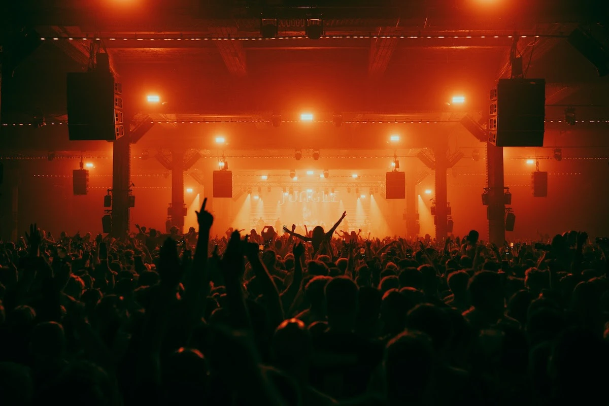 Pista lotada do Warehouse Project, com o público de braços para cima diante do palco, sob luz laranja e fileiras de caixas de som penduradas